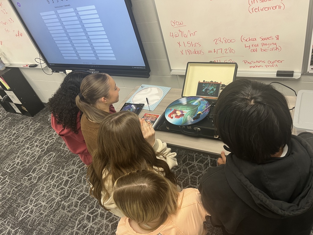 Students gather around a record player in a classroom, examining a colorful vinyl record and working together to play media using older audio technology.
