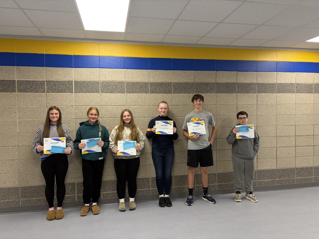 Six Baltic School District students stand together in a school hallway, proudly displaying their September Student of the Month certificates and recognition awards. The students pose in front of a gray brick wall with blue and gold stripes, representing the school's colors.