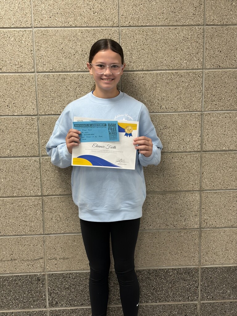 A Baltic School District student wearing a light blue sweatshirt and glasses smiles while holding a September Student of the Month certificate and recognition award. The student stands in front of a gray brick wall inside the school.