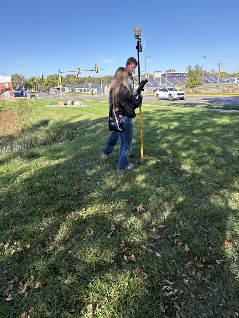 A student works alongside an instructor using GPS surveying equipment mounted on a tall pole during an outdoor hands-on learning activity near a roadway.