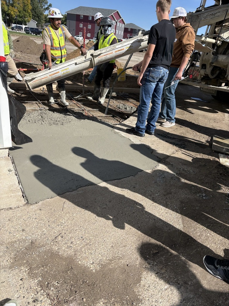 Two students observe construction workers pouring and leveling fresh concrete at an outdoor job site. Workers wearing hard hats and high-visibility safety vests guide concrete from a mixer truck into a prepared area.