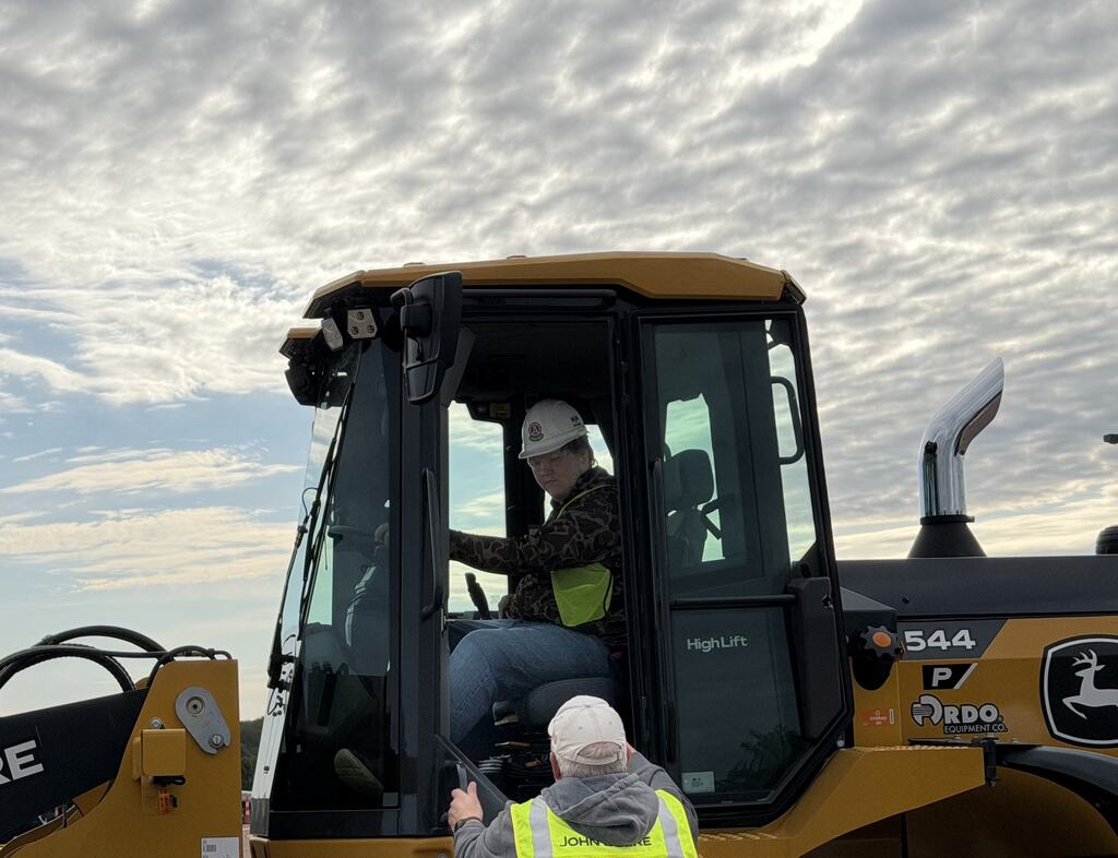 A student wearing a hard hat and high-visibility safety vest sits inside the cab of a John Deere wheel loader while an instructor standing outside provides guidance during a hands-on heavy equipment demonstration.