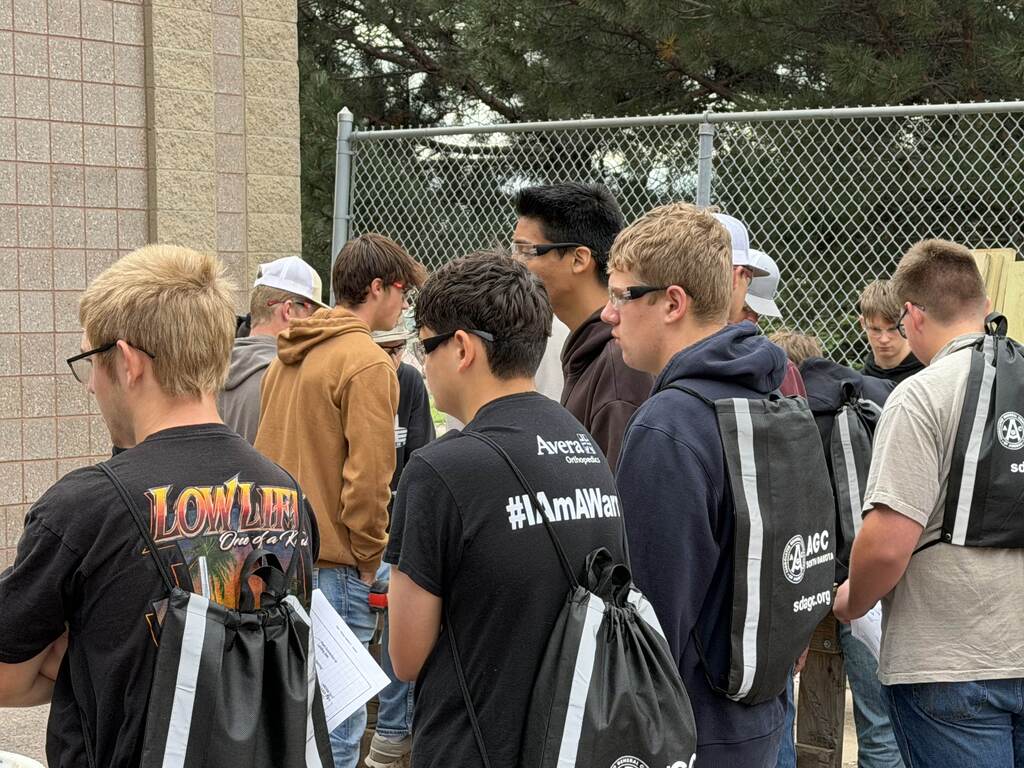 A group of students wearing safety glasses and carrying South Dakota AGC drawstring bags gather outdoors near a fenced training area during a construction and technical education event.