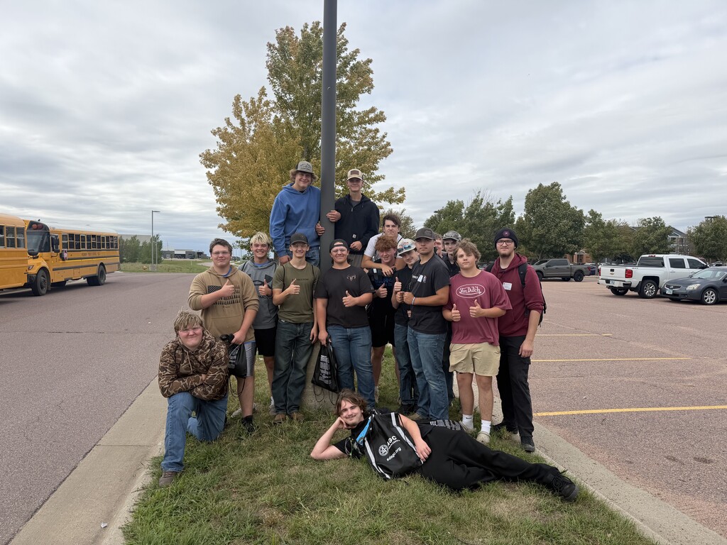 A group of students pose together outdoors near a school bus and parking lot following a technical education event. Several students give thumbs-up gestures while others smile for the camera, including one student reclining playfully on the grass in front of the group.
