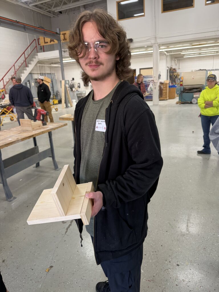 A student stands inside a technical education workshop, proudly displaying a wooden project he constructed. Workbenches, tools, and other participants are visible in the background.