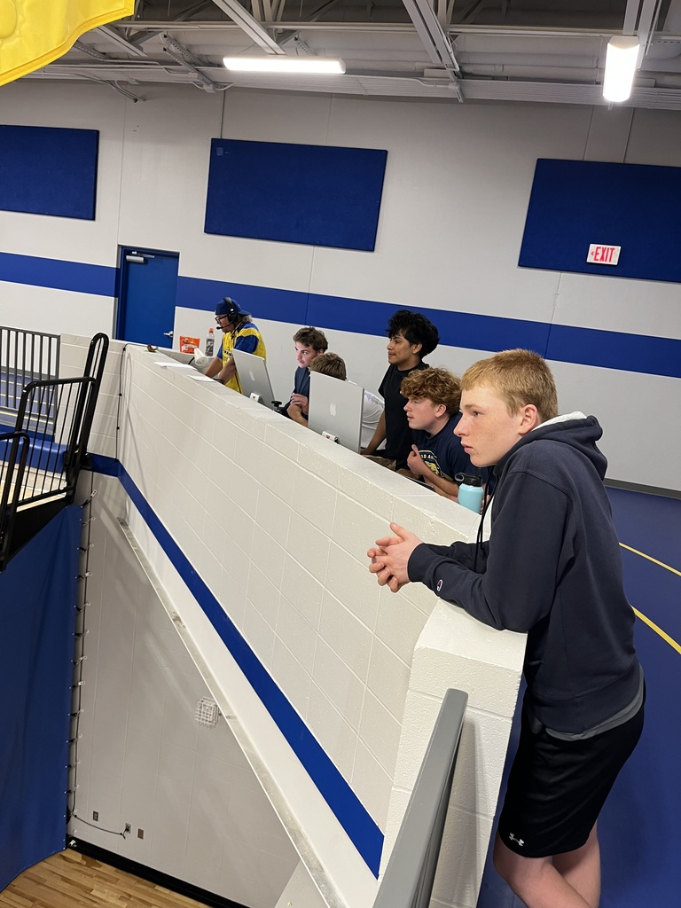 A group of Baltic School District students and an adult gather at the elevated gymnasium media station. Several students operate computers while others watch activities taking place on the gym floor below.