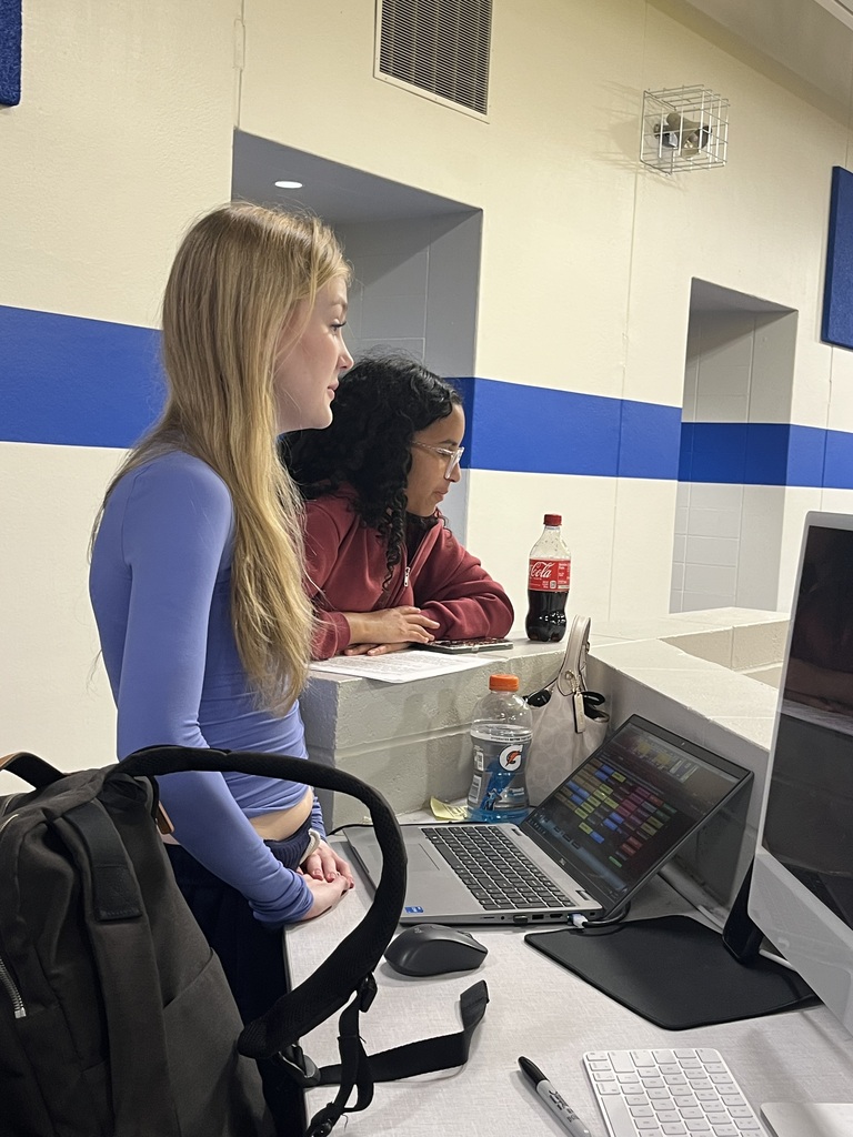 Two students work together at a media production station overlooking the school gymnasium. One student monitors a laptop displaying event production software while the other observes the screen.