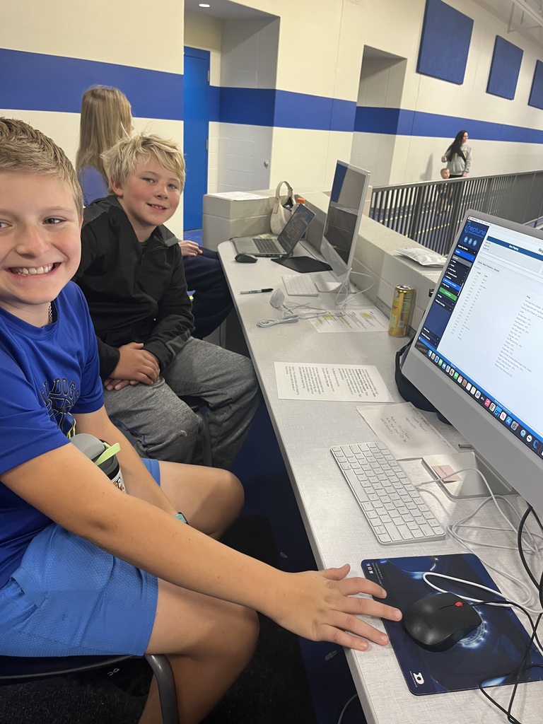 Two Baltic School District students smile while seated at the gymnasium's media and production station. Computer monitors, keyboards, and event management software are visible as students help support school activities.