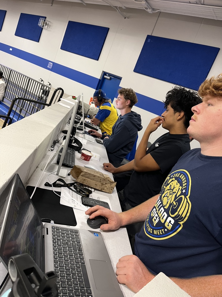 Three students and an adult work together at the gymnasium's media station, operating computers, audio equipment, and event production software during a school activity.