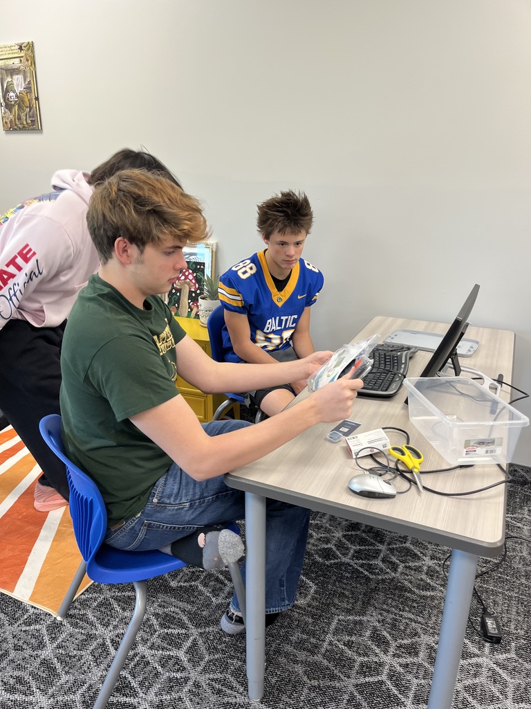 Three students collaborate around a computer workstation during a Raspberry Pi project. One student examines the project materials while another watches the computer and a third observes the activity.