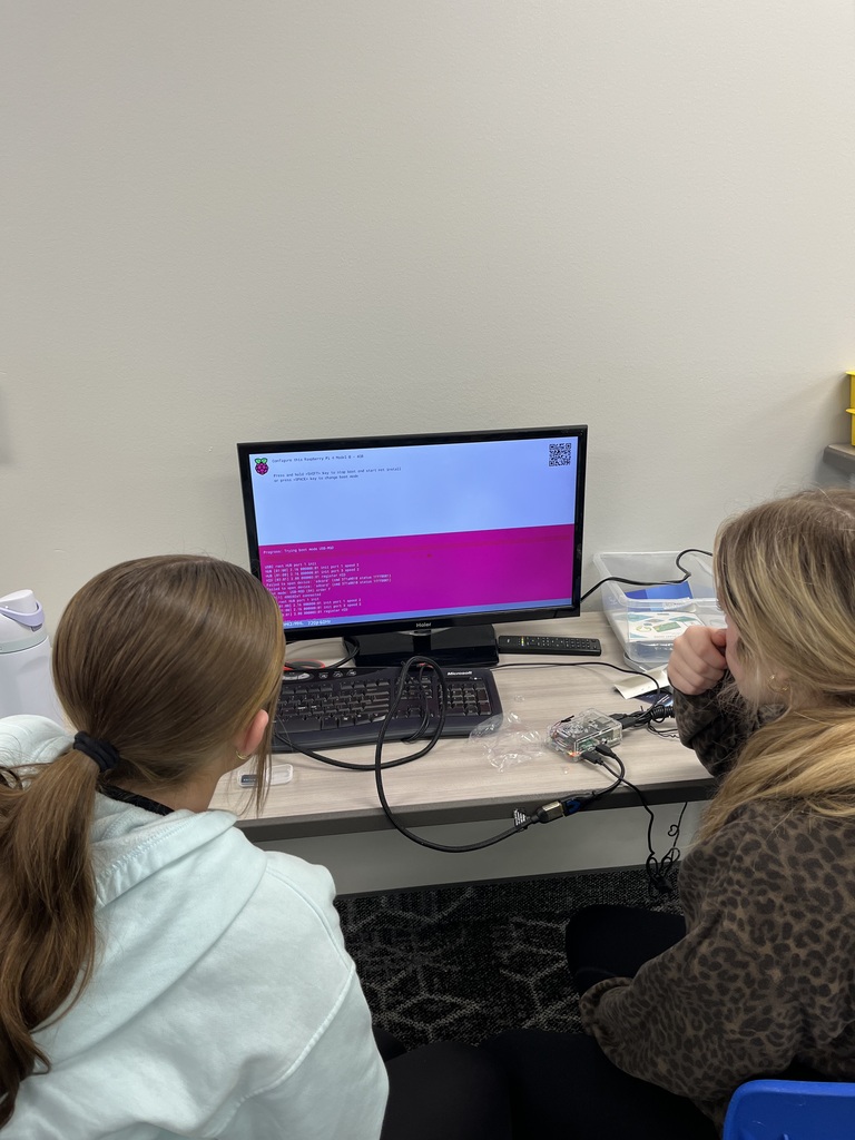 Two students work together at a computer station, observing a monitor displaying the Raspberry Pi operating system installation and setup process.