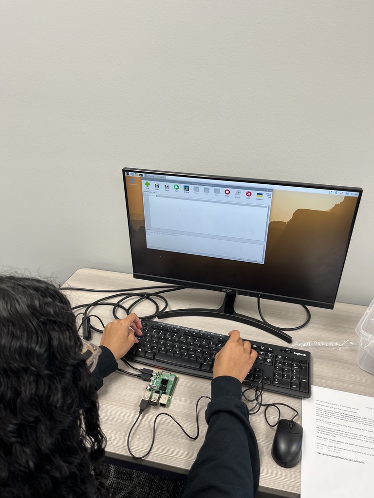 A student uses a keyboard to write Python code on a computer connected to a Raspberry Pi single-board computer during a classroom programming activity.