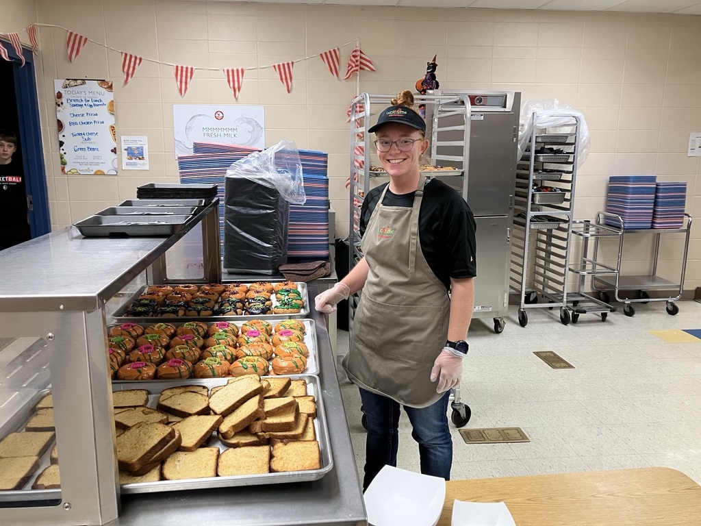 A smiling Lunchtime Solutions food service employee stands beside trays of Halloween-themed donuts and breakfast items in the Baltic School District kitchen, preparing for the Donuts with Grownups event.