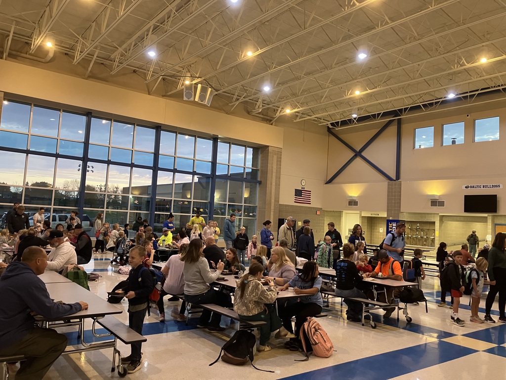 Students, parents, and family members gather in the Baltic School District commons to enjoy breakfast together during the Donuts with Grownups event. Families sit at cafeteria tables, visit, and share a morning meal.