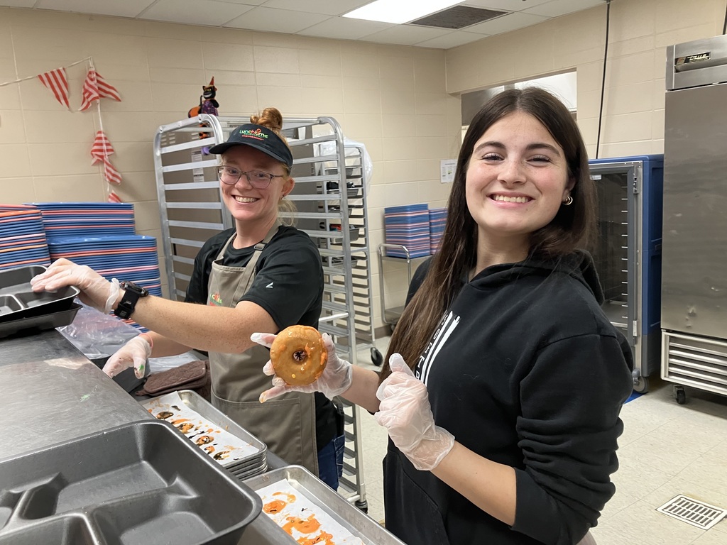 A Lunchtime Solutions food service employee and another smiling helper pose in the school kitchen. One holds a Halloween-themed donut while both wear food service gloves and help prepare breakfast for the Donuts with Grownups event.