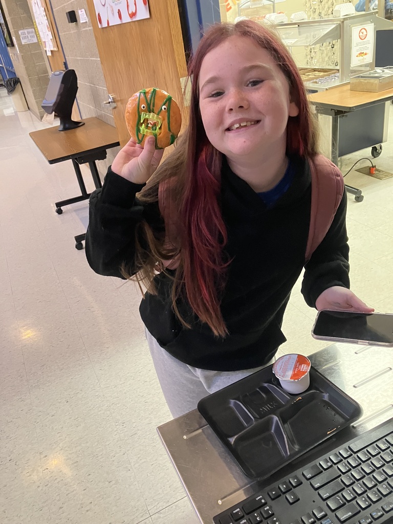 A smiling student holds up an orange-frosted Halloween donut decorated with green icing and candy teeth while standing near the school cafeteria.