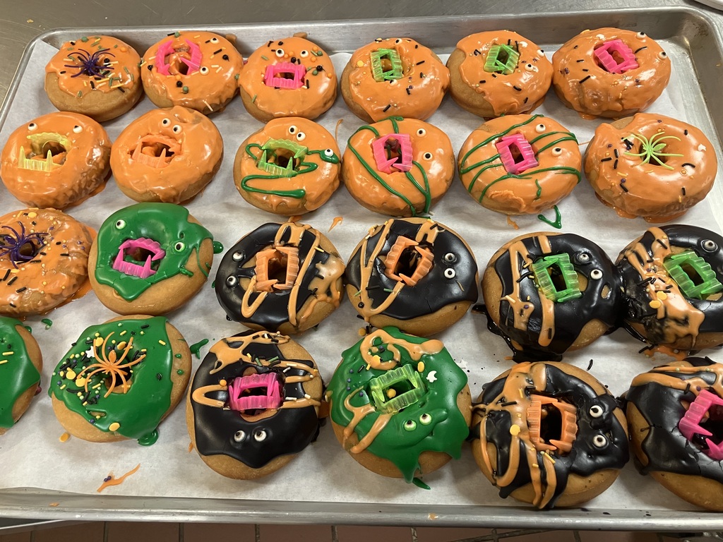 A tray of Halloween-themed donuts decorated with orange, green, and black frosting, candy eyeballs, colorful sprinkles, plastic vampire teeth, and spiders, prepared for the Donuts with Grownups breakfast event.