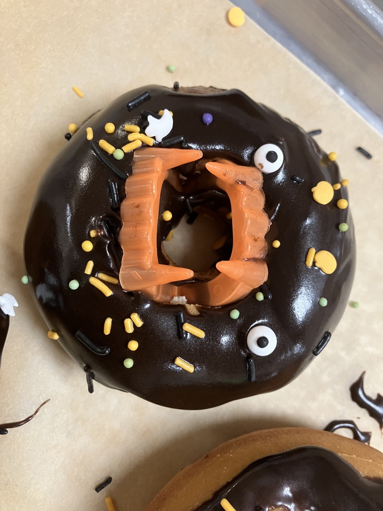 Close-up of a Halloween-themed donut covered in dark chocolate frosting, decorated with orange vampire teeth, candy eyeballs, and colorful sprinkles.