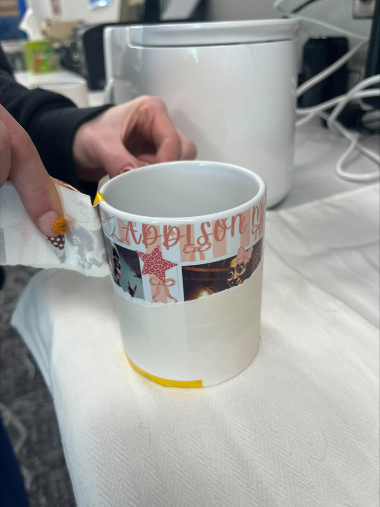 Close-up of a white ceramic mug featuring a personalized pink and red printed design with the name “Addison.” A student carefully peels away the transfer paper to reveal the finished artwork.
