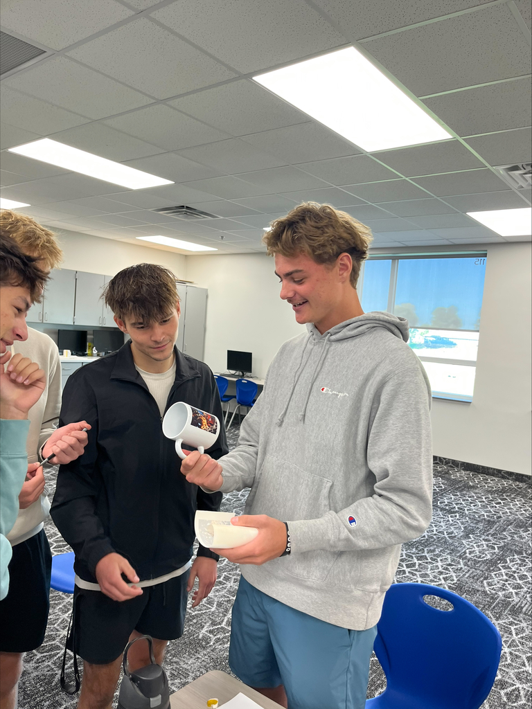 A group of students gather around a classmate who displays a white ceramic mug featuring a colorful printed design during a hands-on crafting activity.