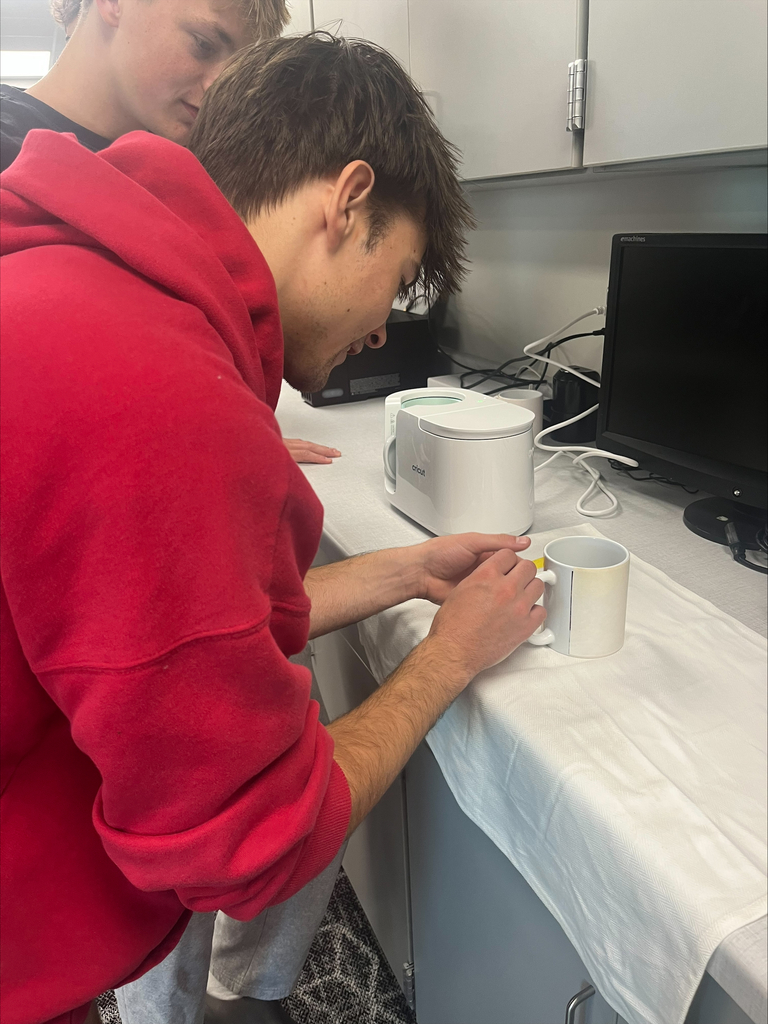 Two students collaborate at a classroom workstation, securing a printed design onto a white ceramic mug using heat-resistant tape before transferring the design.