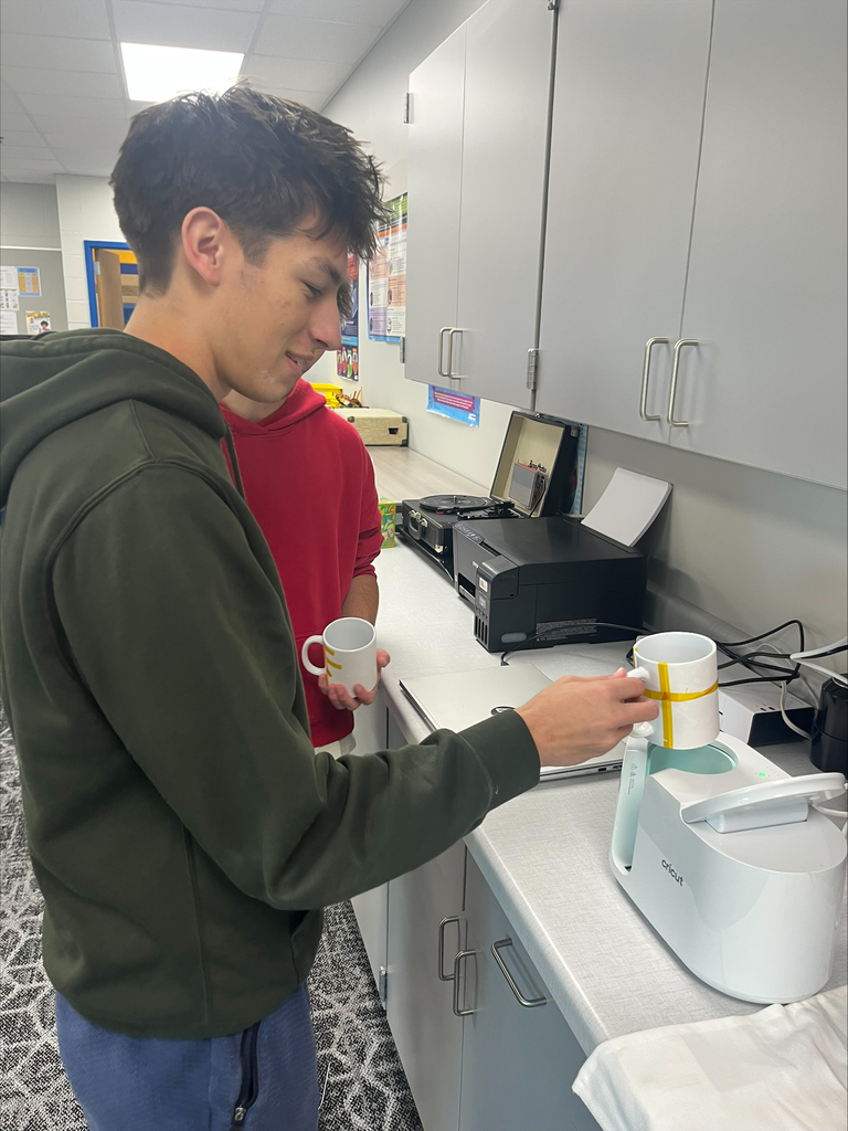 Two students work together at a Cricut mug press, placing a ceramic mug with a taped design into the machine to transfer a custom graphic.