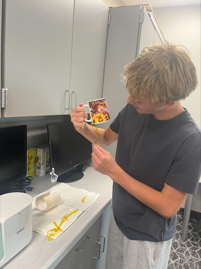A student carefully removes yellow heat-resistant tape from a custom-printed ceramic mug while working at a classroom counter near a Cricut mug press.