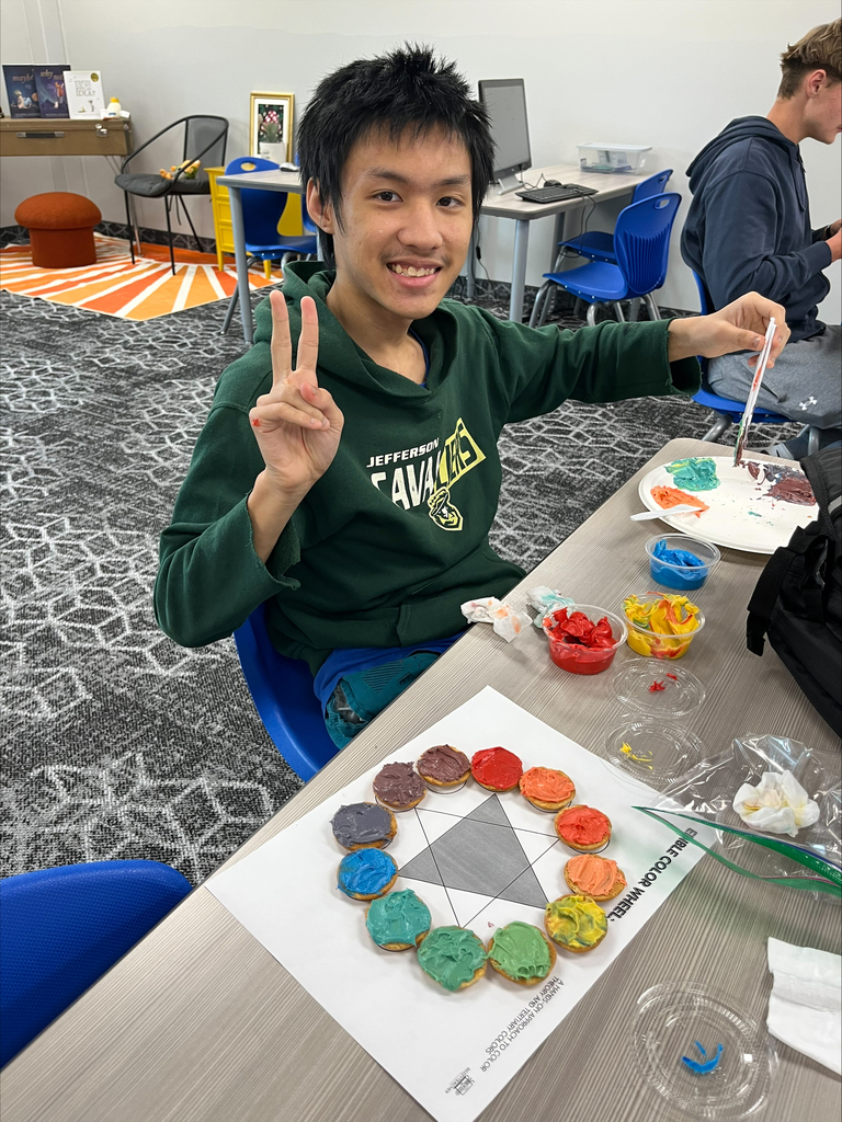 A student wearing a green sweatshirt smiles and holds up a peace sign beside a completed edible color wheel. Cookies decorated with colorful frosting are arranged in a circle to demonstrate primary, secondary, and tertiary colors.