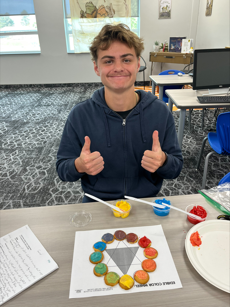 A Baltic School District student wearing a navy blue sweatshirt smiles and gives two thumbs up while displaying a completed edible color wheel made from cookies decorated with different colors of frosting.
