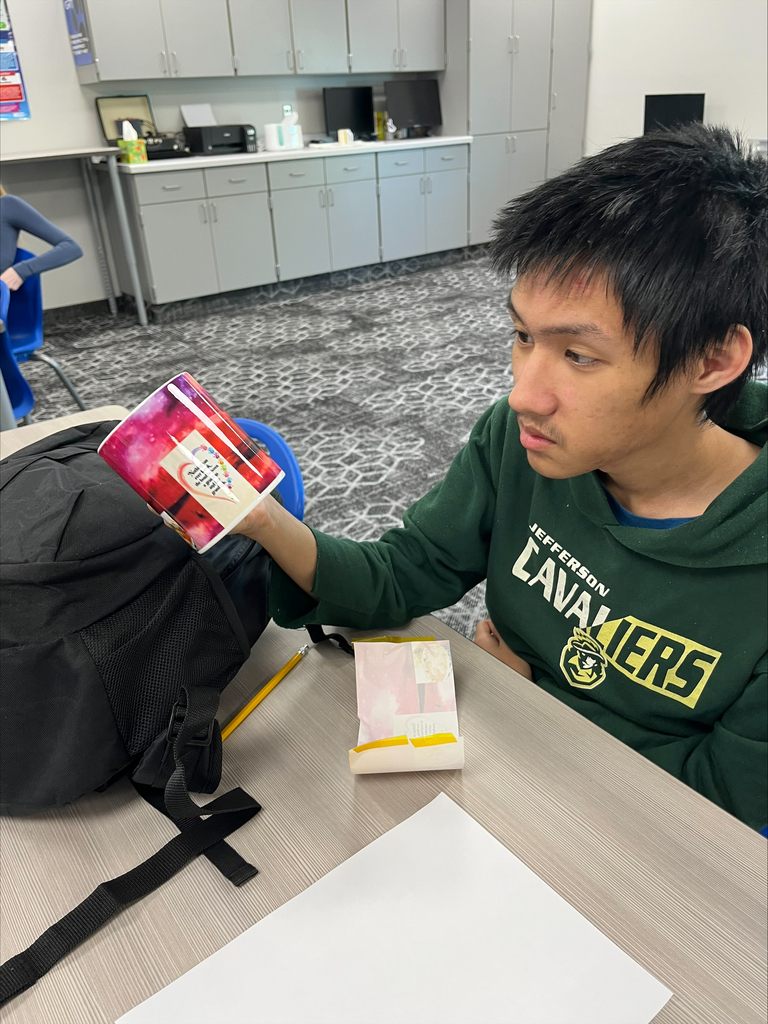 A student wearing a green Jefferson Cavaliers sweatshirt examines a custom-designed ceramic mug with a red and pink printed graphic during a classroom crafting project.