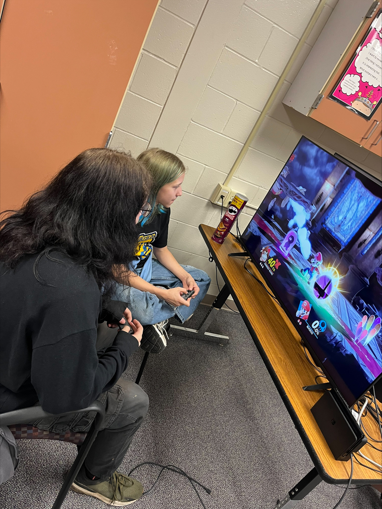Two students sit side by side holding Nintendo Switch controllers while playing Super Smash Bros. Ultimate on a television during an E-Sports activity.
