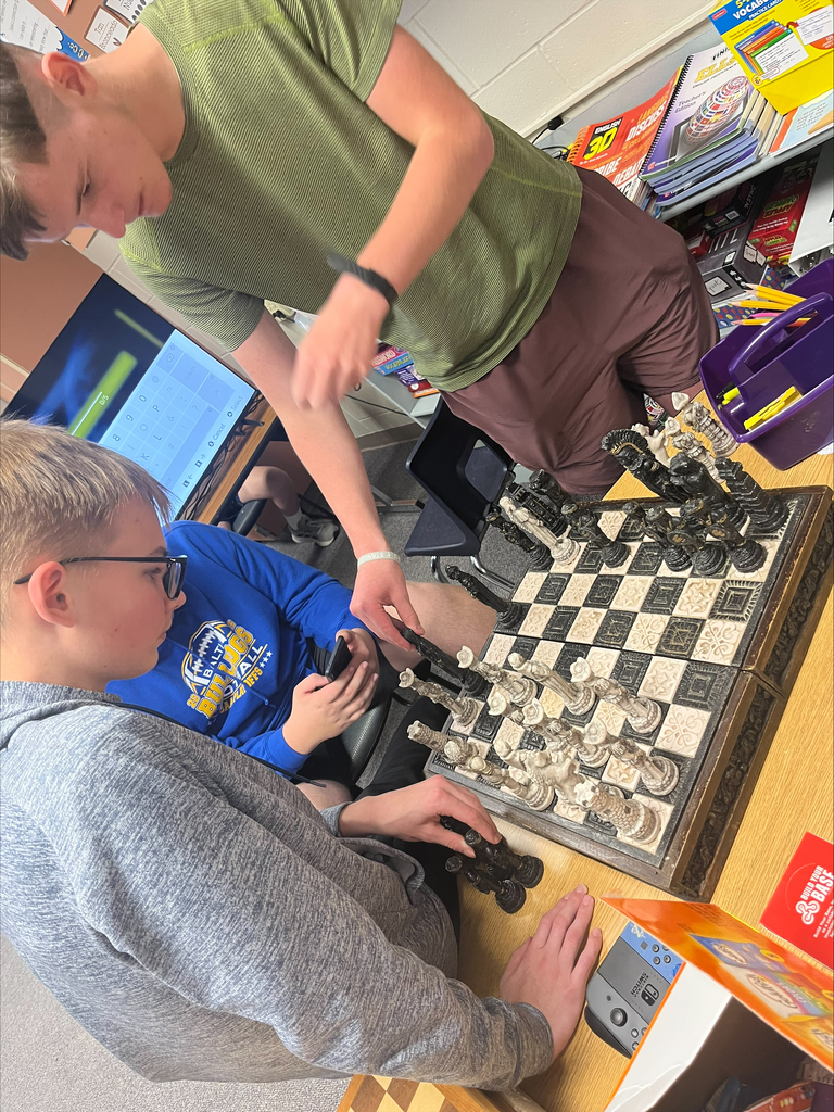 Three students gather around a decorative chessboard, discussing moves and examining the pieces during a strategy-based activity.
