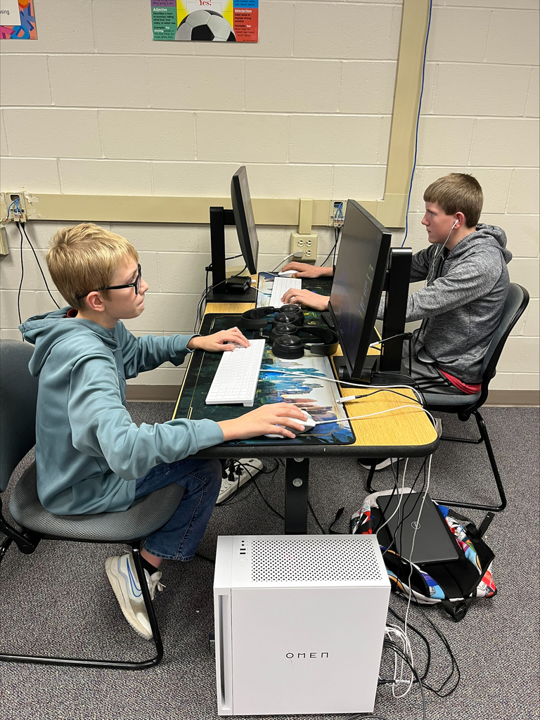 Two students sit at desktop gaming computers, using keyboards and mice during an E-Sports activity. Gaming headsets and a white OMEN gaming computer are visible at their workstation.