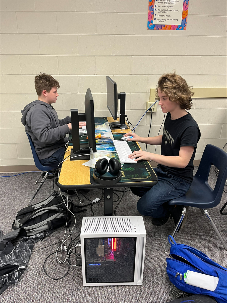 Two students focus on their individual gaming monitors while using keyboards and mice. A gaming computer with illuminated internal components sits beneath their shared desk.