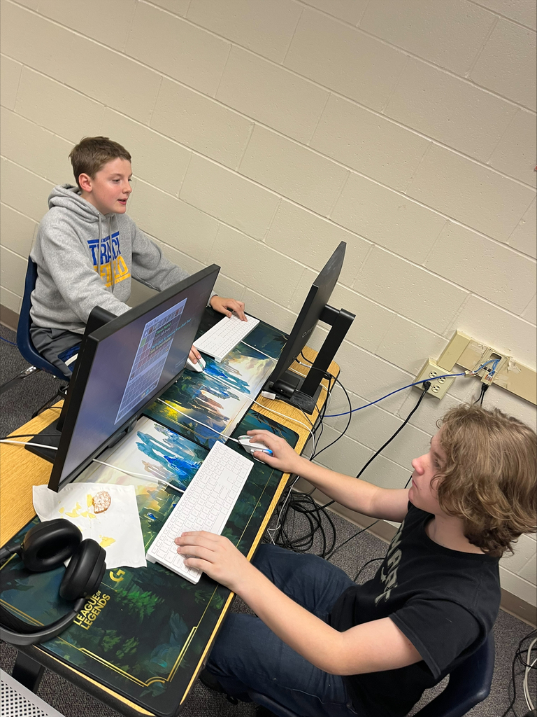 Two students compete at adjacent gaming computers while a third student sits nearby. One monitor displays Minecraft gameplay during the E-Sports session.