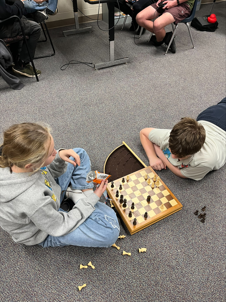 Two students sit on the classroom floor playing chess on a wooden board, with captured pieces scattered nearby.