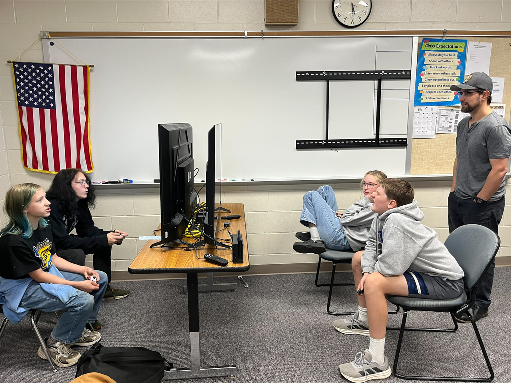 Four students participate in a multiplayer video game session using controllers and a dual-monitor setup while an adult observes. The students sit across from one another in a classroom.