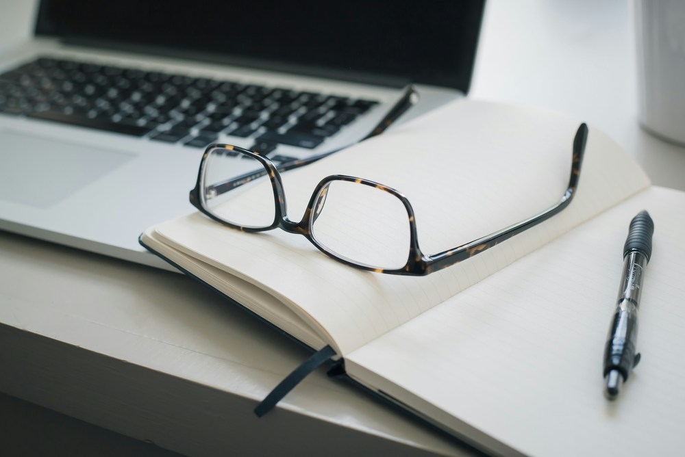 glasses laying on a notebook next to a pen on a desk with a laptop