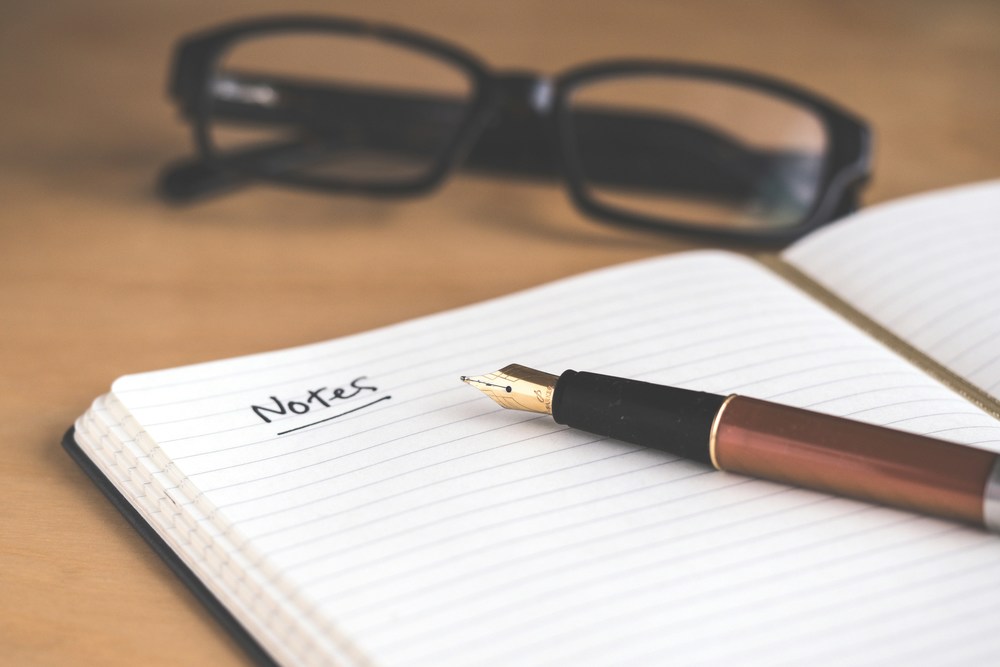 notebook laying on a desk with a pen on top and glasses laying nearby