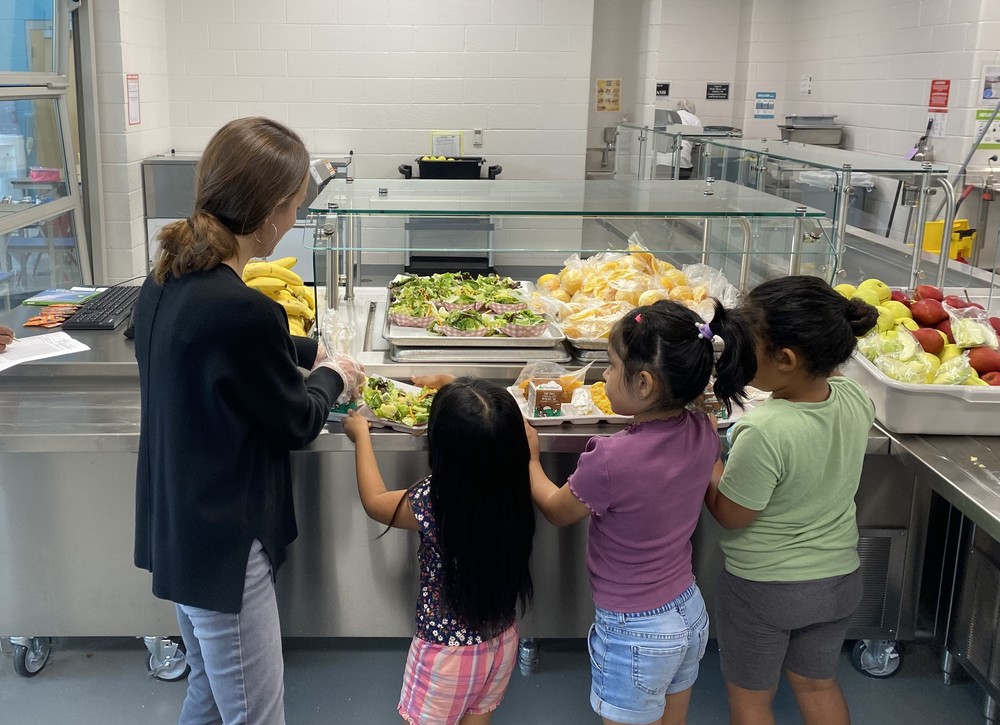 woman putting food on trays for three young girls in the cafeteria