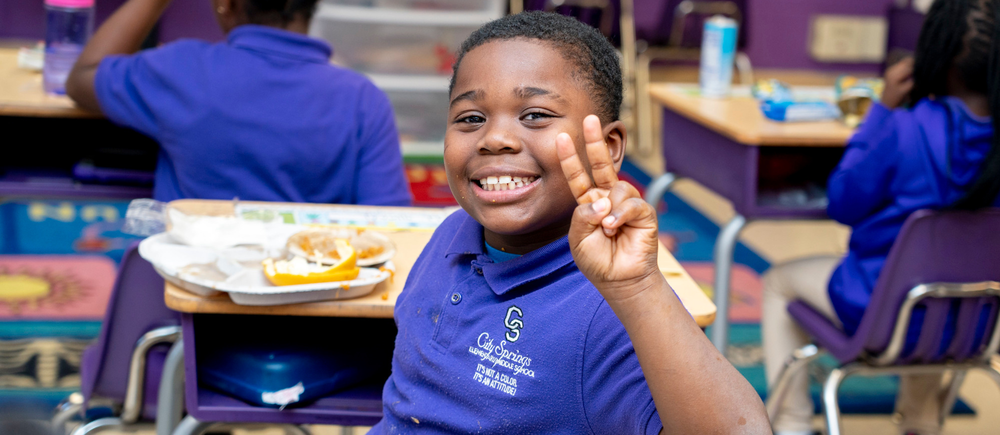 Smiling boy in a blue school uniform showing a peace sign while sitting at a classroom desk with a meal tray.