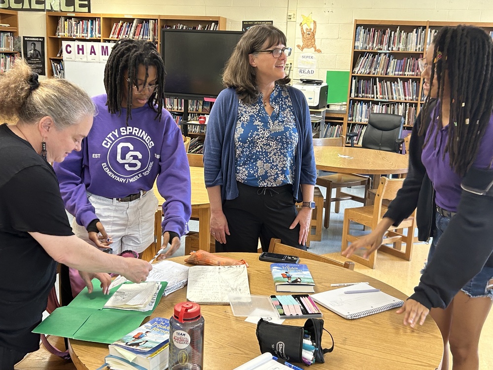 Students, teachers, and Kate Messner gathered around a library table, chatting and looking at schoolwork.