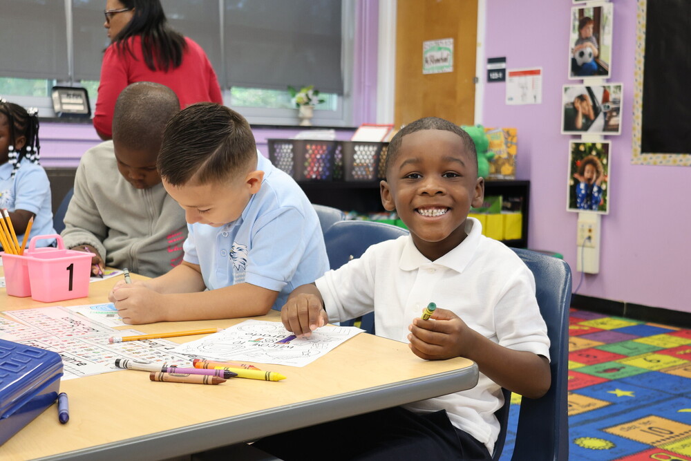 Students coloring at their desk. One students smiles at the camera.