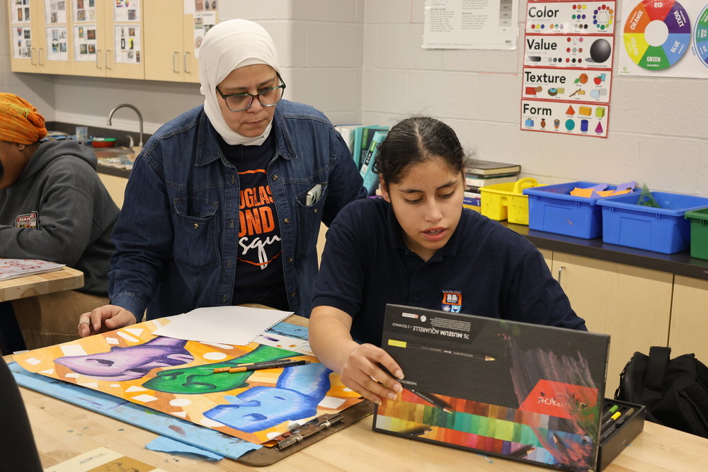 Art teacher assists student with chalk drawing on paper during class.