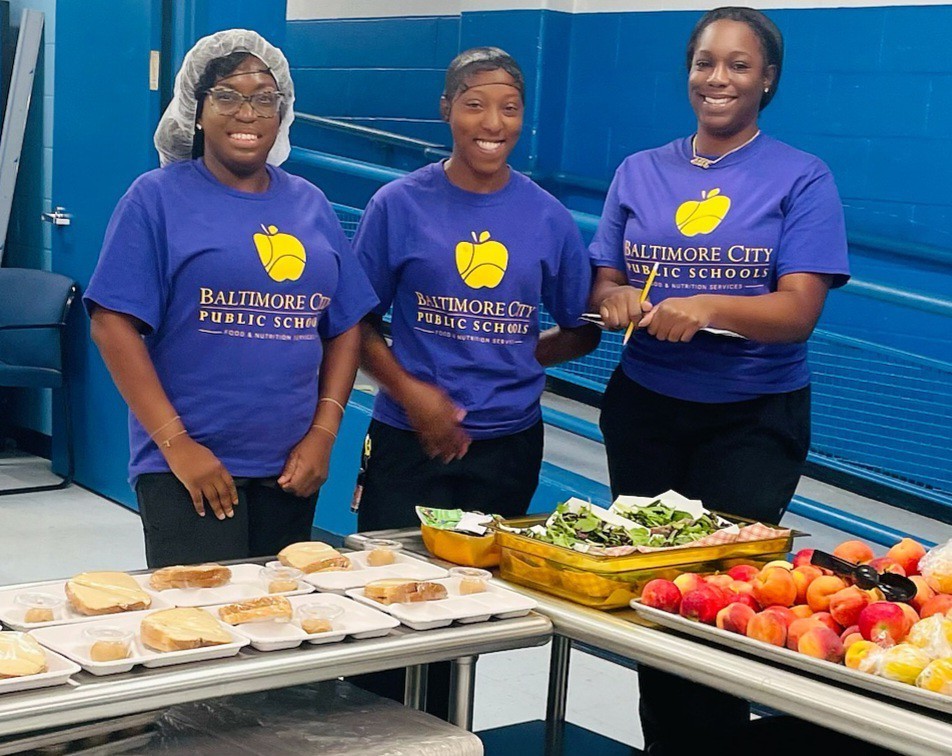 Food service staff members line up to take photo smiling, all wearing matching shirts to celebrate the first day of school. 