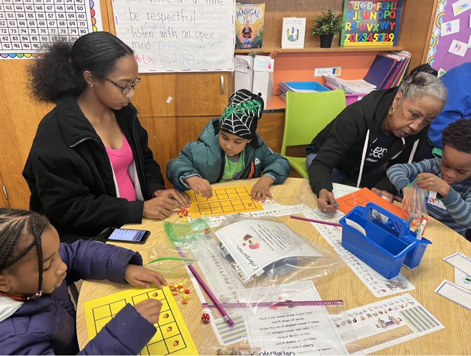 Two women and two young students are sitting at a table completing a learning worksheet