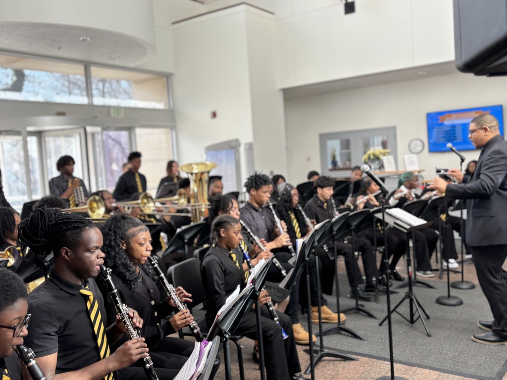 The Reginald F. Lewis band in black shirts/pants with yellow and black striped ties performing with their instruments in the lobby at Central Office
