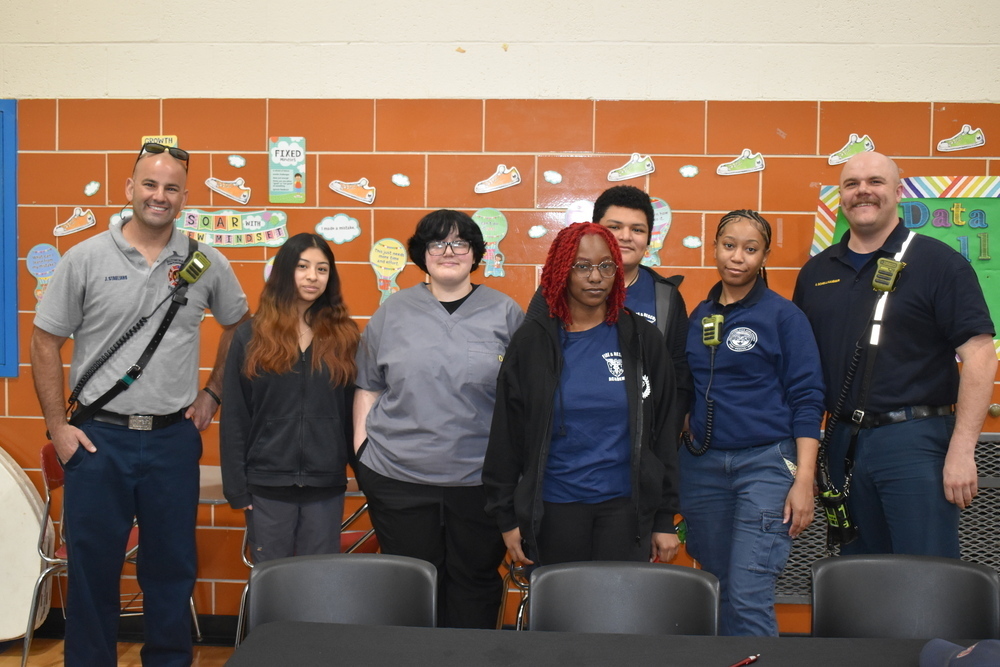 Group photo of two uniformed officers and five diverse young adults standing in front of a classroom bulletin board with colorful decorations.