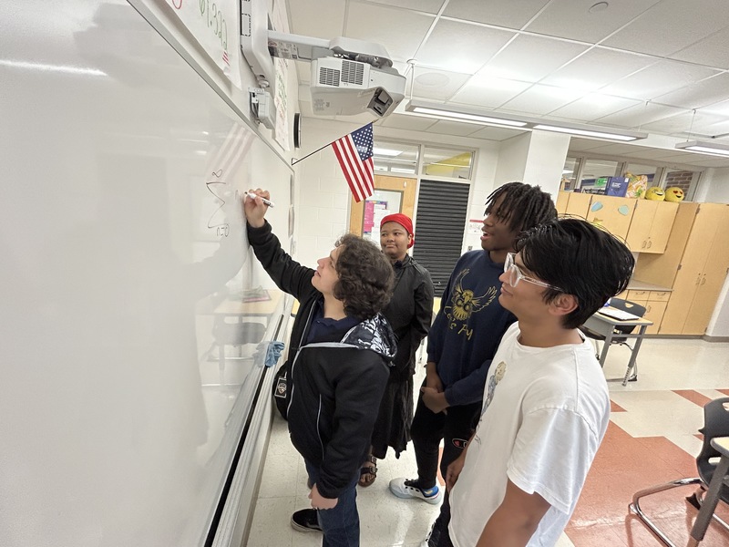 Sudarshan, Jordan, Oscar, and Audrey go over a math problem on the whiteboard of their classroom. 