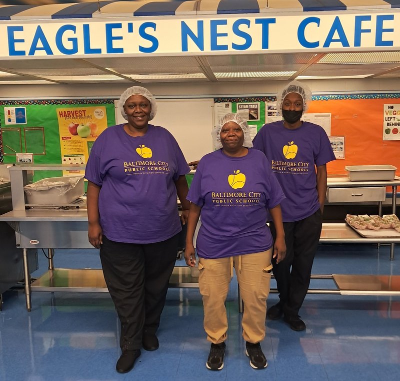Food service staff members line up to take photo smiling in the cafeteria, all wearing matching shirts to celebrate the first day of school. 